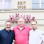 Luis Dias, Klaus Höhn und Cyril Becker stehen vor dem alten Höhns Gebäude in Köln mit dem Schild "Höhns" und "Seit 1908"