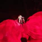 Helene Fischer in der LANXESS arena, performend in einem riesigen roten Kleid, das mithilfe von Luft und Seilen in Form gehalten wird, während sie mit einem Mikrofon vor dem Mund singt.