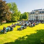 Klassische Bugatti-Autos auf einer Wiese vor einem eleganten Herrenhaus ausgestellt, mit Teilnehmern, die sich unterhalten und die Oldtimer bewundern.