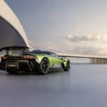 Rückansicht eines grünen und schwarzen Aston Martin Valhalla Supersportwagens mit ausgefahrenem Heckflügel auf einer hellen, ebenen Fläche vor einer geschwungenen Wand und einem langen Steg unter klarem Himmel.