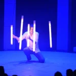 Ein Künstler auf der Bühne des GOP Varieté-Theaters Bonn kniet hinter leuchtenden Stäben, die von der Decke hängen, während er in blaues Licht getaucht ist.